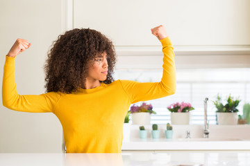 African american woman wearing yellow sweater at kitchen showing arms muscles smiling proud. Fitness concept.
