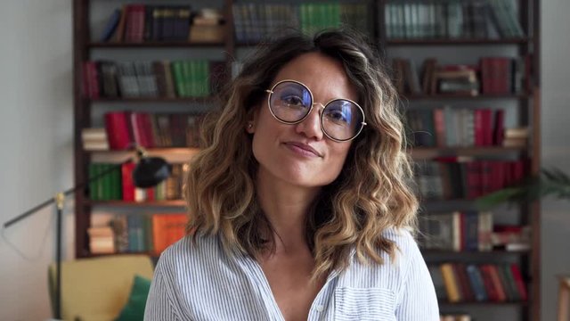 Beautiful curly woman in thirties of fair complexion in white casual shirt and round fashionable glasses looks softly into camera and smiles openly and positively in spacious room with large bookcase