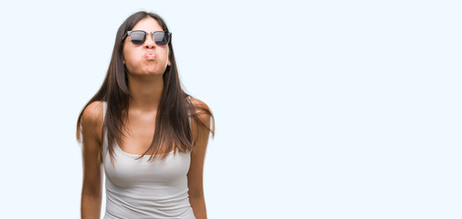 Young beautiful hispanic wearing sunglasses puffing cheeks with funny face. Mouth inflated with...