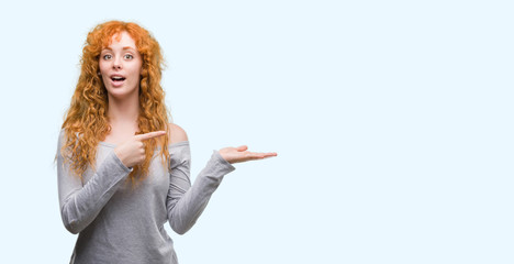 Young redhead woman amazed and smiling to the camera while presenting with hand and pointing with...
