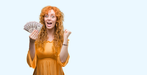 Young redhead woman holding dollars screaming proud and celebrating victory and success very excited, cheering emotion
