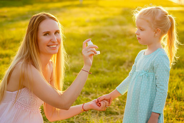 beautiful and young smiling woman holding child by the hand and gives inhaler white against park...