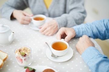 Fototapeta na wymiar Hands of senior couple by table with cups of drinks, strawberry dessert in glass and white porcelain teapot during tea time