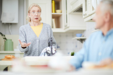 Anxious blond mature woman with towel talking to her husband in the kitchen