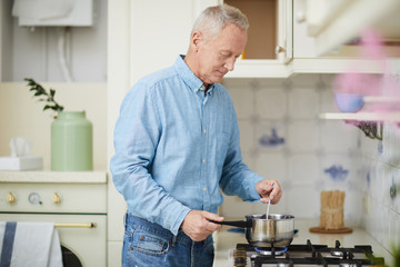 Senior man mixing something in pan while standing by gas stove and cooking - obrazy, fototapety, plakaty