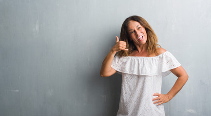 Middle age hispanic woman standing over grey grunge wall smiling doing phone gesture with hand and fingers like talking on the telephone. Communicating concepts.