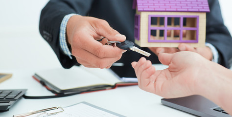Real estate agent in suit holds house model in one hand and in the second hand holds out the keys of the house to woman.