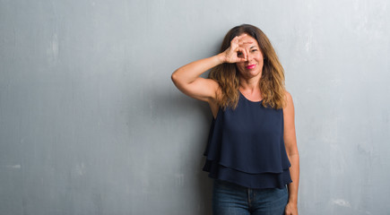 Middle age hispanic woman standing over grey grunge wall doing ok gesture with hand smiling, eye looking through fingers with happy face.