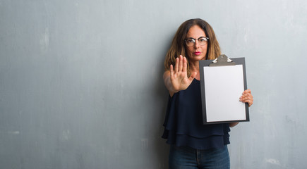 Middle age hispanic woman standing over grey grunge wall holding clipboard with open hand doing stop sign with serious and confident expression, defense gesture