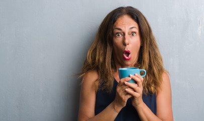 Middle age hispanic woman standing over grey grunge wall drinking cup of coffee scared in shock with a surprise face, afraid and excited with fear expression
