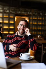 Happy businessman in casualwear and eyeglasses talking by smartphone while relaxing in cafe