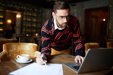 Busy young employee leaning over table while making notes and looking through online data in laptop