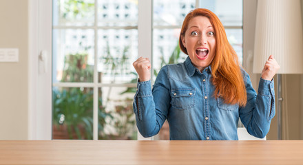 Redhead woman at home celebrating surprised and amazed for success with arms raised and open eyes. Winner concept.