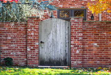 Rustic wooden arched garden fence in brick wall in autumn with colored leaves and brick house with colorful fall foliage reflected in windows