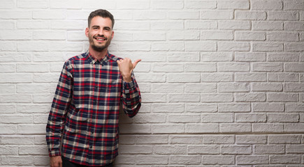 Young adult man over white brick wall pointing and showing with thumb up to the side with happy face smiling