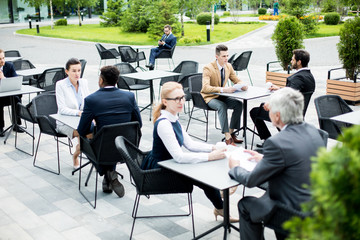 Small groups of employees in formalwear sitting by tables in outdoor summer cafe, relaxing and...