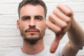 Obraz premium Young adult man standing over white brick wall with angry face, negative sign showing dislike with thumbs down, rejection concept
