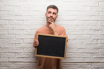 Young adult man over brick wall holding blackboard serious face thinking about question, very confused idea