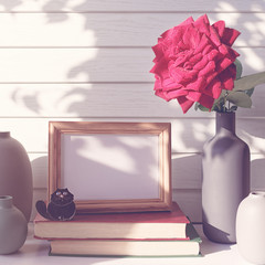 A fresh red rose with drops of water on the petals is in a gray vase. Nearby is a stack of books, a photo frame and a black cat figure. White wooden background