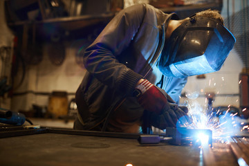 Young welder in protective mask leaning over workbench and welding iron workpieces