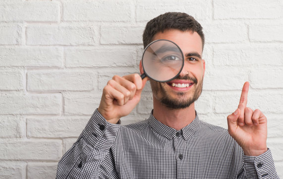 Young adult man over brick wall using magnifying glass surprised with an idea or question pointing finger with happy face, number one