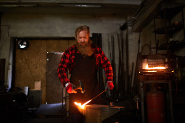 Fototapeta na wymiar Bearded blacksmith in flannel and apron standing by anvil and forging hot molten metal