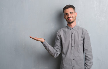Young adult man standing over grey grunge wall smiling cheerful presenting and pointing with palm...