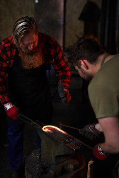 Two men in workwear processing molten metal workpiece with handtools on anvil