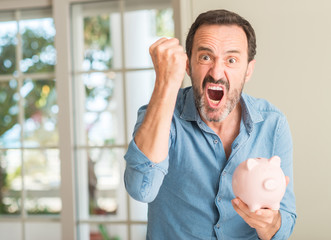 Middle age man save money on piggy bank annoyed and frustrated shouting with anger, crazy and...