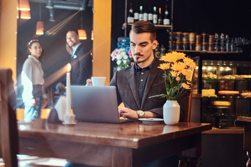 Handsome freelancer man with stylish beard and hair dressed in a black suit working on laptop while sitting at a cafe.