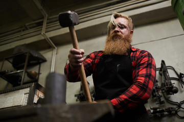 Blacksmith with beard holding big hammer over anvil while forging iron workpiece in smithy © pressmaster