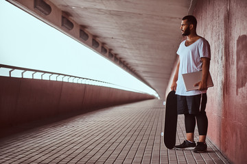 African-American bearded guy dressed in a white shirt and sports shorts holds on a laptop and...