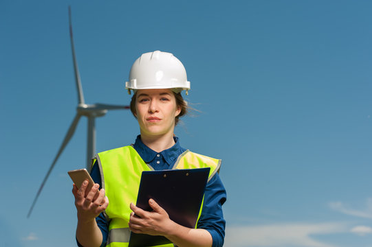 Portrait of a beautiful positive woman engineer in a green vest shirt with a smartphone in hand against the backdrop of a windmill