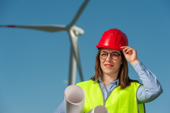 Portrait of a beautiful young female engineer in red helmet and glasses with projects and plans against the background of a windmill and blue sky