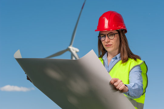 Serious young female engineer in red helmet and glasses with projects and plans against the background of a windmill and blue sky