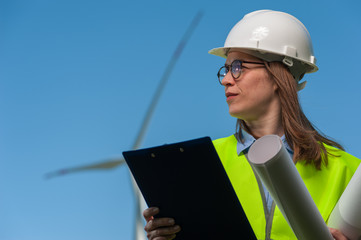 Portrait of a successful young female architect in a safety helmet with a work plan and projects on a background of windmills and blue sky.