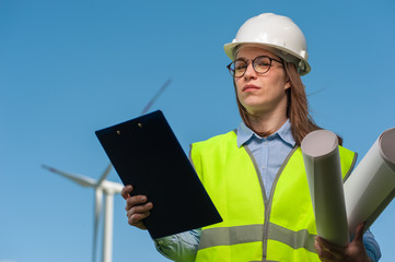 Portrait of a successful young woman with a female engineer in a safety helmet with a work plan and projects on a background of windmills and blue sky.