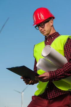 Portrait of a ambitious young male engineer in a safety helmet with a work plan and projects on a background of windmills and blue sky.