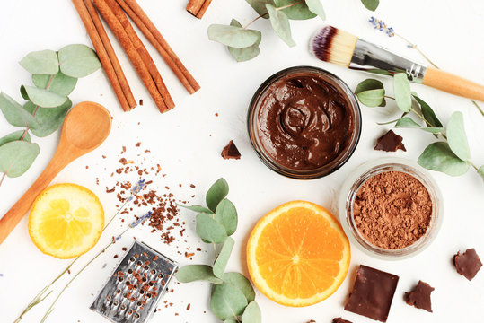 Chocolate skincare mask. Natural ingredients for making beauty treatment products. Cacao, orange slices, aroma oil, jars and spoons, herbal leaves. Top view white table. 