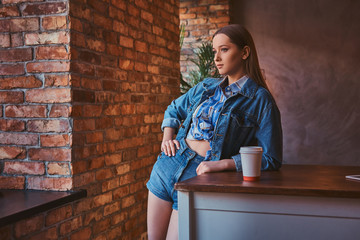 Young hipster girl dressed in a shirt and jeans jacket and shorts leaning on the table with a cup of takeaway coffee in a room with loft interior.