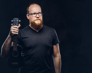 Redhead bearded male musician wearing glasses dressed in a gray t-shirt holds ukulele. Isolated on a dark textured background.