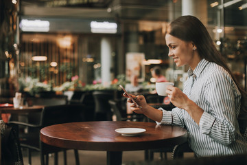 Nice and young girl is sitting at table and looking at phone. She is holding it and smiling. Also girl has a cup of coffee in other hand. She has lunch.
