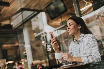 Dark-haired and beautiful woman is sitting at the table and looking at phone. She is smiling. Girl has cup of cofffee on right hand. She is sitting in restaurant.