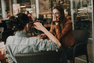Emotional girl is brown jacket is sitting in front of her friend. She is laughing and covering...