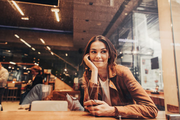 Adorable girl with brown hair sitting at table and looking to the right. She is holding phone in one hand and the other one is close to face. She is dreaming