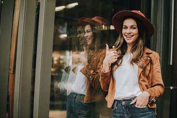 Positive and happy girl standing and posing on camera. She is leaning to glass wall. Girl showing big thumb up. Woman is smiling. The other hand is in pocket