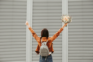 Girl is standing face to wall and holding hands up. She has flowers in hand. Girl wears brown jacket and bag on back. Isolated on striped and white background