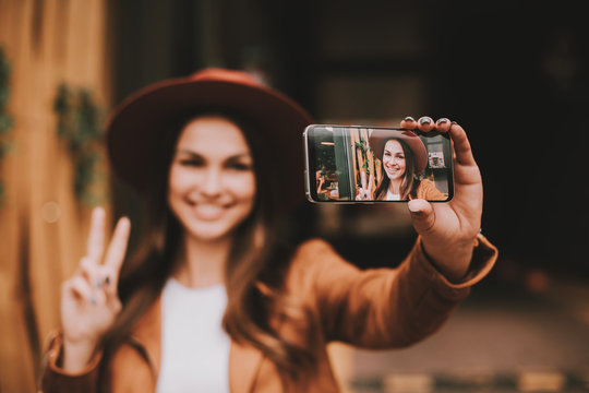 Amazing and positive young woman is standing ans posing on camera. She is taking selfie. Girl smiling and showing piece symvol with fingers. Woman is happy