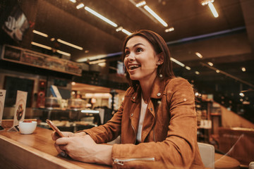 Excited and happy woman looking forward. She is amazed. Girl is sitting and holding phone in hands. She is in restaurant
