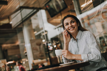 Portrait of business woman sitting at table in cafe. She is talking on the phone and smiling. Girl is looking to the side. She is holding cup of coffee in roght hand. Woman looks happy